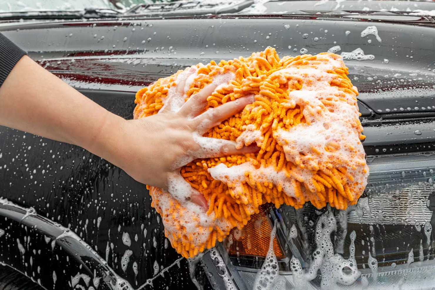 Person washing a car with an orange sponge covered in soap suds.